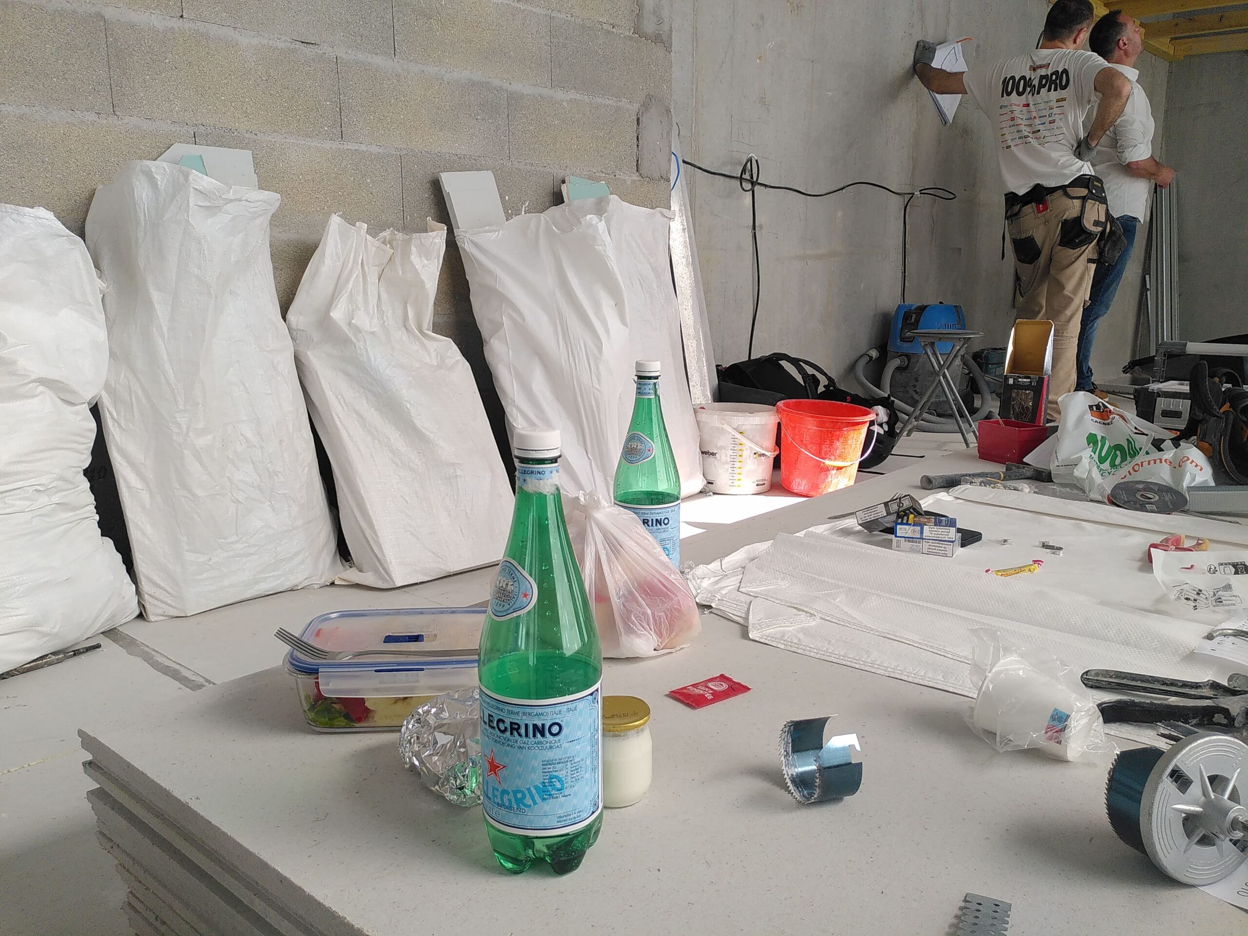 Dans l'atelier d'auto-réparation vélo de Paris 10 Paillettes et Cambouis, des sacs sont entreprosés contre un mur de béton brut, des ouvriers discutent, au premier plan on voit des bouteilles en plastique.